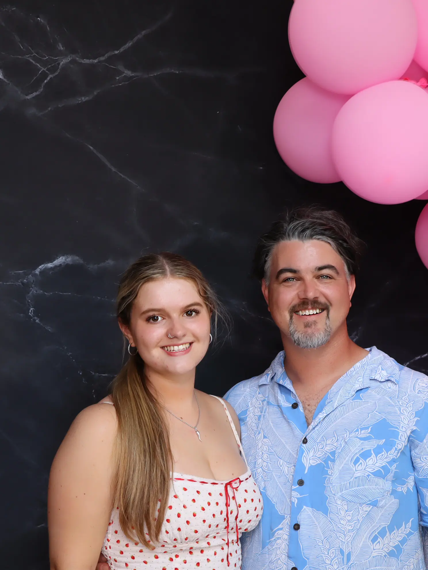A smiling couple poses under pink balloons against a dark backdrop.
